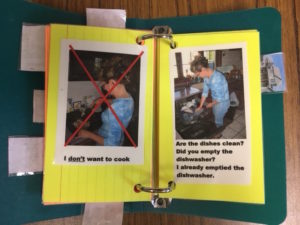 The photo depicts a communication book page of a woman placing dishes in the dishwasher along with printed sentences including the following: 1) Are the dishes clean? 2) Did you empty the dishwasher? and 3) I already emptied the dishwasher. 