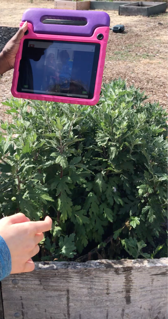 A child's left hand is holding up an iPad showing a video of gardening. The child is standing in front of a large planter.