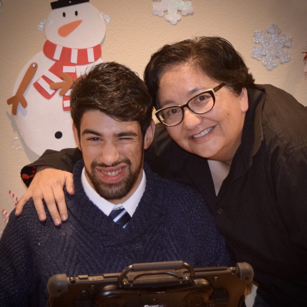 Karen stands with arm around Cristian who is sitting with an AAC device.