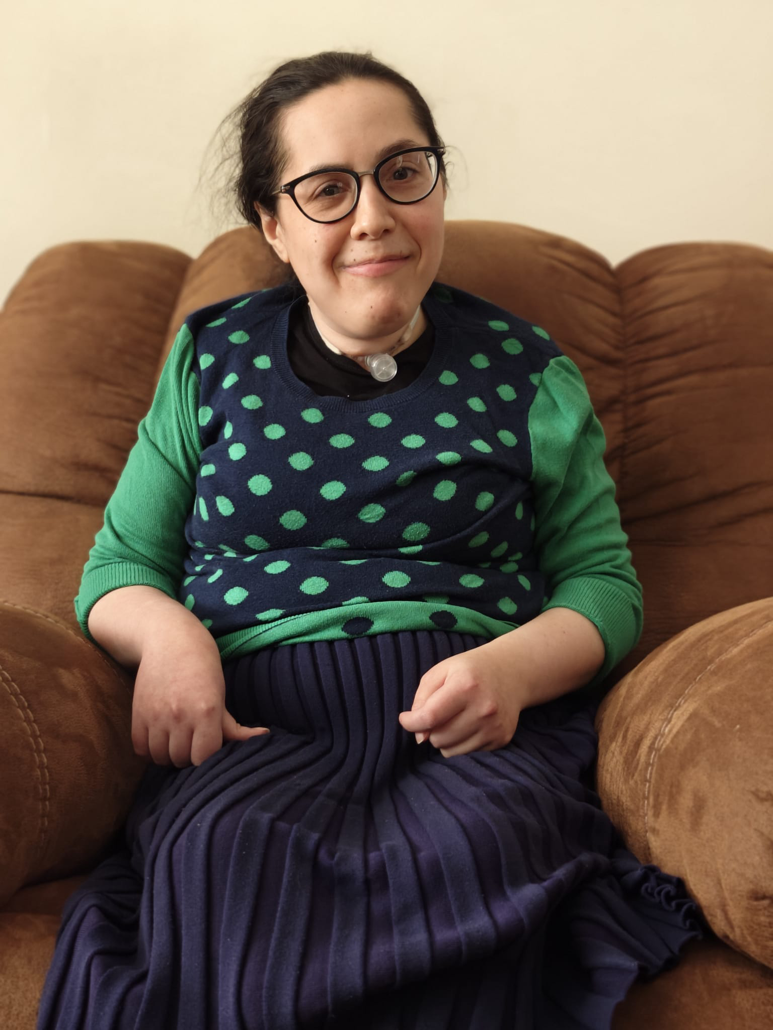 Esther, a woman wearing glasses and a tracheostomy, sits on a couch and smiles at the camera.